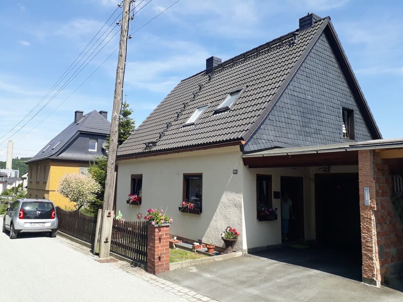 Two-story house with slate roof and flower boxes on a sunny street next to a parked car.
