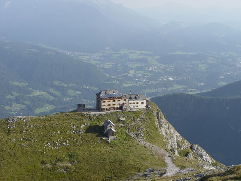 Watzmannhaus mountain hut perched on a grassy cliff edge overlooking a hazy valley with distant towns.