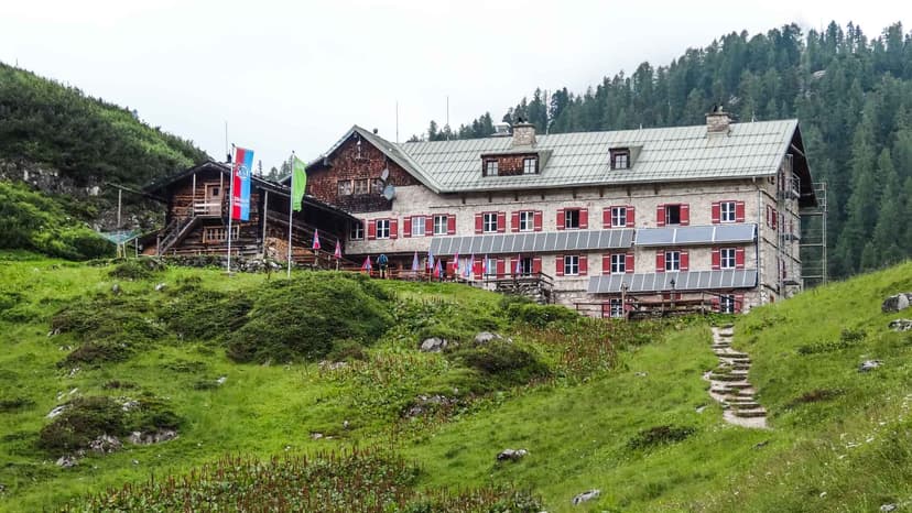 Stone mountain hut with red shutters and solar panels on grassy slope near forest