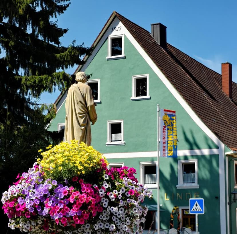 Statue in front of green Gasthof Linde building with colorful flower basket and tall pine tree.