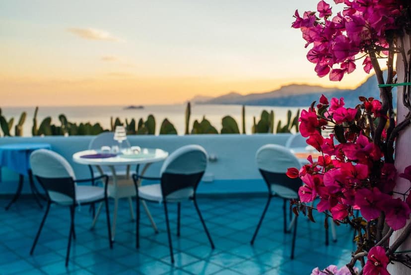 Outdoor dining terrace with pink bougainvillea overlooking the sea and mountains at sunset.