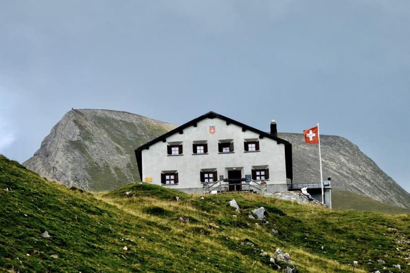 Alpine hut with Swiss flag on grassy slope below rocky mountain peak under gray sky