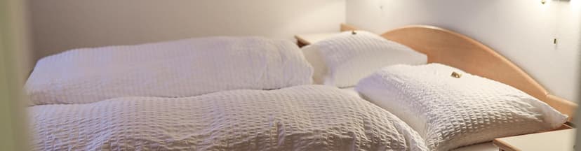Hotel room bed with white textured linens and pillows, light wood headboard.