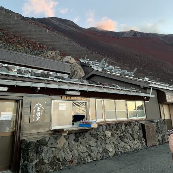 Mountain hut with stone base against dark volcanic slope, Munatsuki Sanso sign visible.