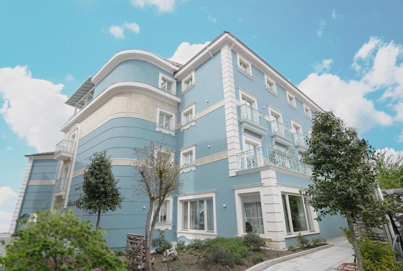 Boutique hotel exterior with light blue facade, white trim, and small trees under a cloudy sky.