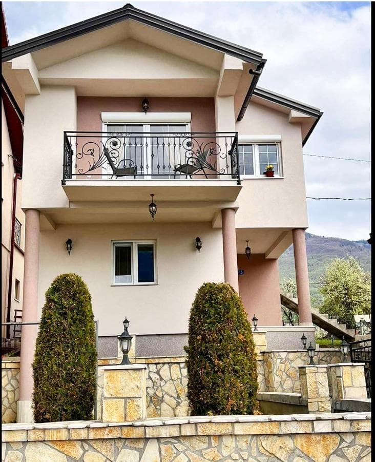 Two-story apartment building with stone retaining wall and manicured bushes, mountains in background.