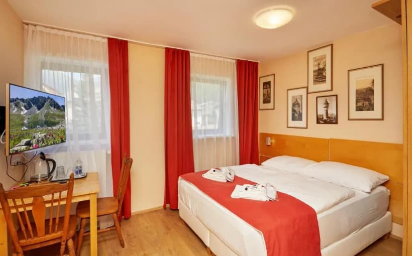 Hotel room with double bed, desk, TV showing mountains, and framed art on the wall.