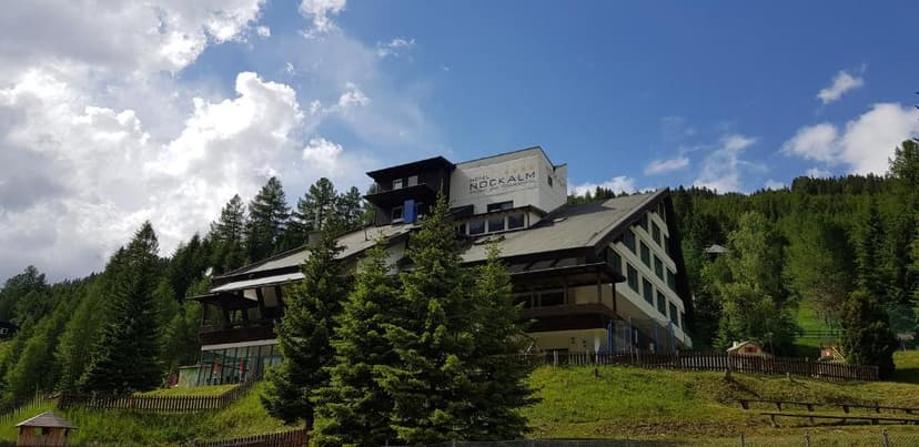 Hotel Nockalm building nestled on a grassy slope surrounded by dense pine forest under a partly cloudy sky.