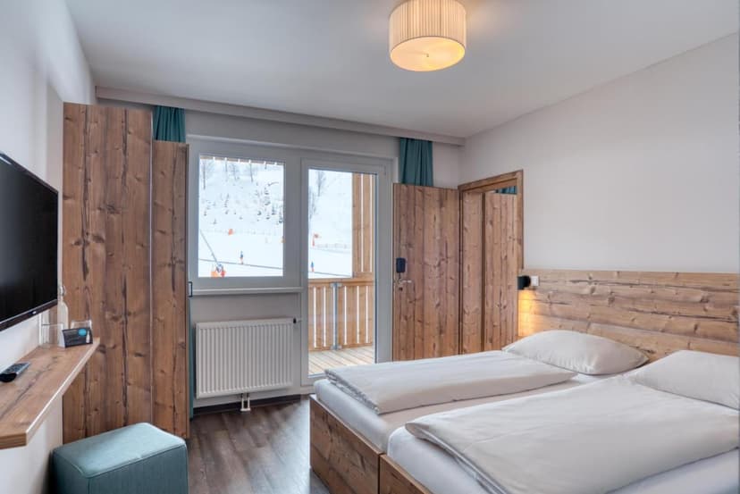 Hotel room with twin beds, wood paneling, and balcony view of snowy ski slope in Bad Kleinkirchheim.