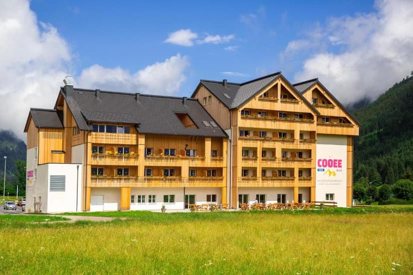 COOEE alpin Hotel Dachstein building with wooden balconies in a grassy field against mountains.
