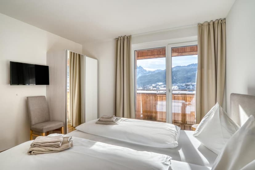 Hotel room with twin beds, wall-mounted TV, and balcony view of snowy Dachstein mountains.