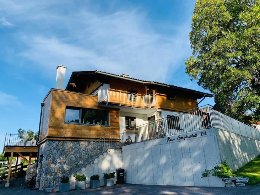 Haus Bergkristall 191 building with wood siding, stone base, and metal railings under blue sky.