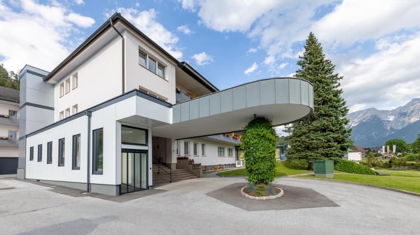 Hotel Grimmingblick entrance with modern canopy, green lawn, and alpine mountains in background.