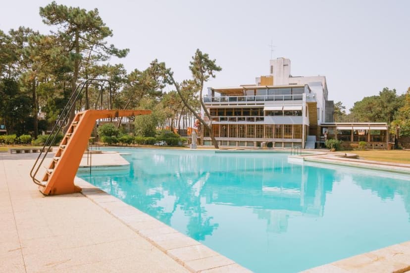 Orange diving board next to a large outdoor pool with a modern building and pine trees.