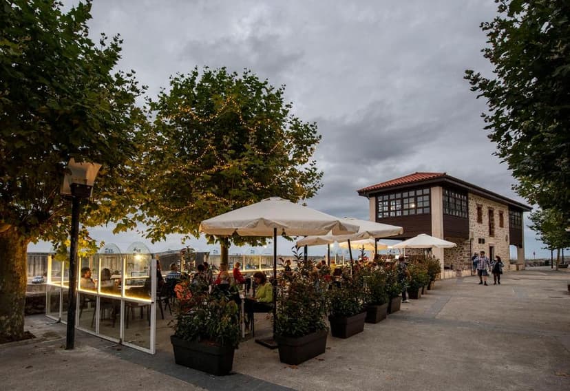 Outdoor dining patio with umbrellas, string lights in trees, and stone building under cloudy sky