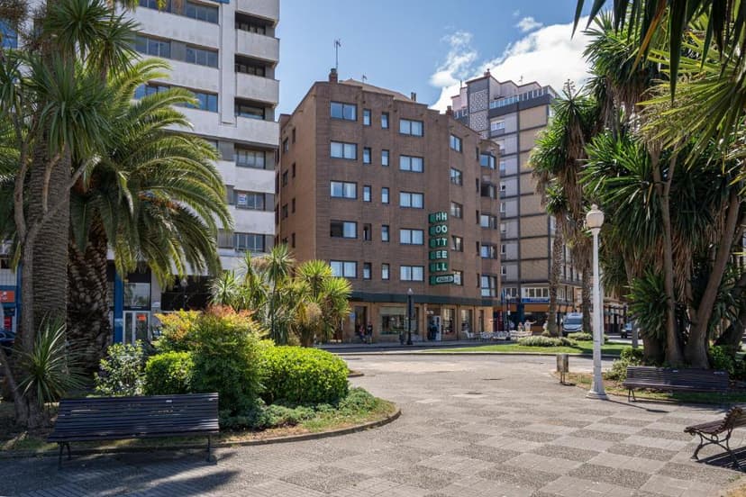 Hotel Alda Pasaje building facade seen from a sunny public square with palm trees and benches.