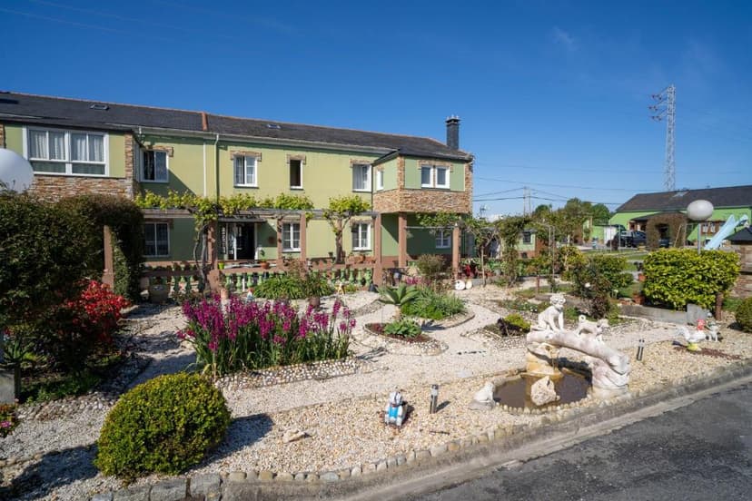Green house with elaborate gravel garden, purple flowers, and stone statues under blue sky.