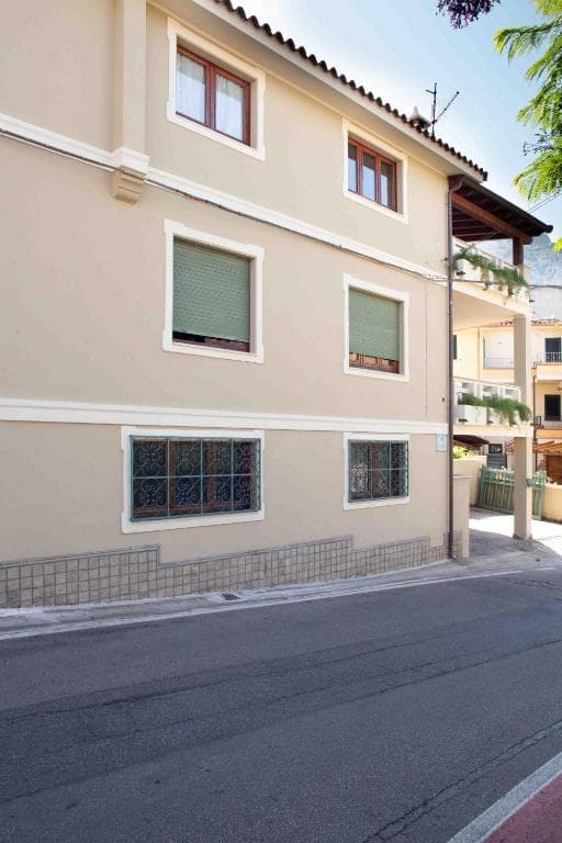 Two-story beige building with green-shuttered windows and wrought iron grates on a street in Casa Pilighitta.
