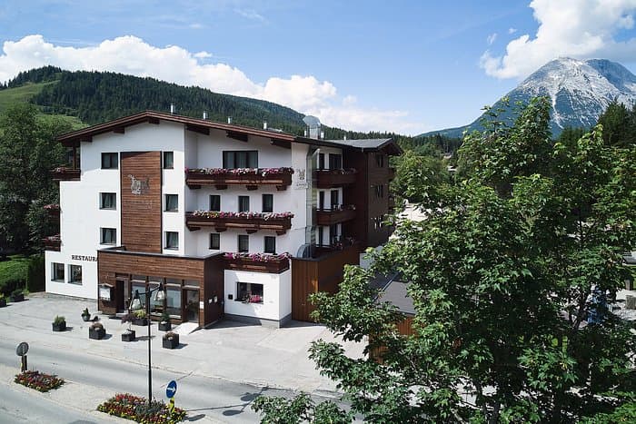 Alpine hotel with wooden balconies and restaurant in front of a large, snow-capped mountain.