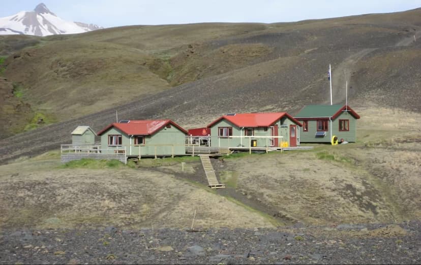 Green huts with red roofs nestled in barren, rolling hills below a snow-capped peak.
