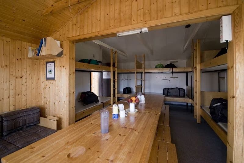 Interior of a wooden mountain hut with bunk beds, a long wooden table, and a seating area.