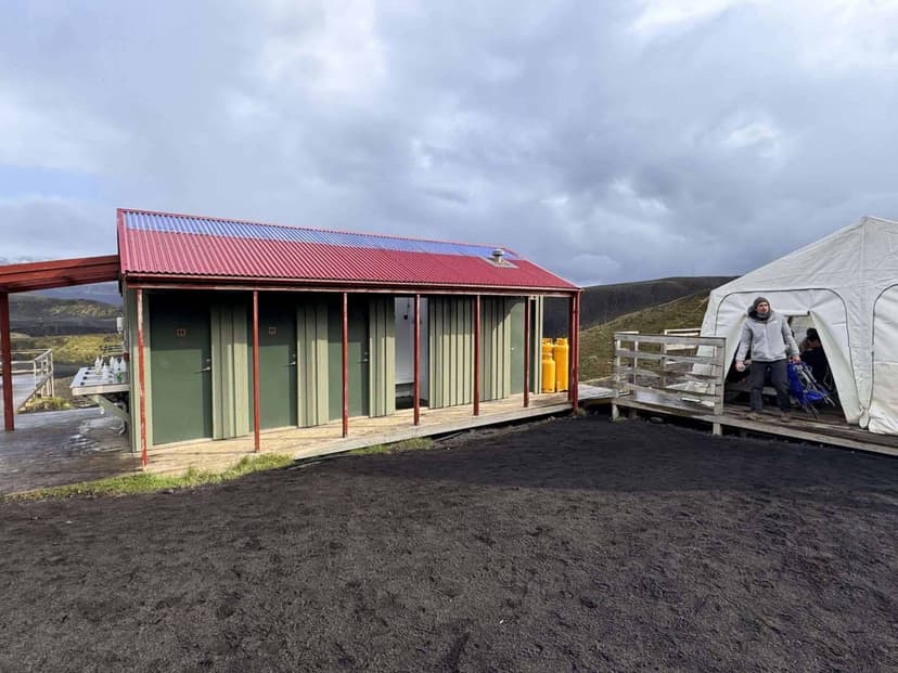 Hut with red roof, white tent, and person standing on black volcanic ground under cloudy sky.