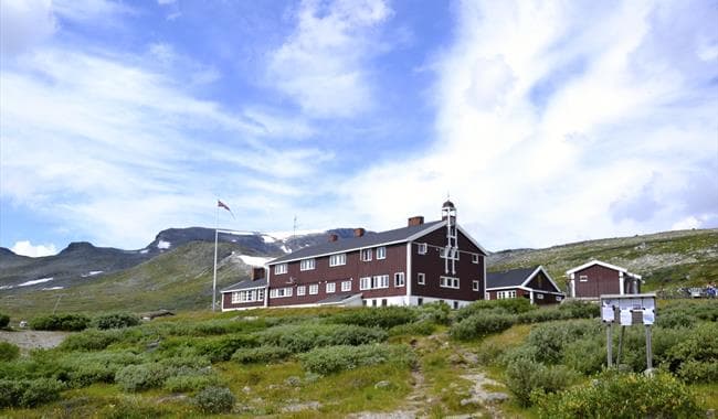 Glitterheim lodge in a green mountain landscape with snow patches under a cloudy blue sky.