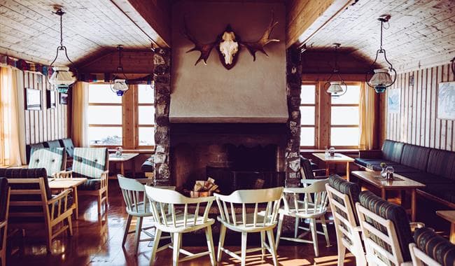 Lodge interior with stone fireplace, antler mount, wooden chairs, and bright windows.