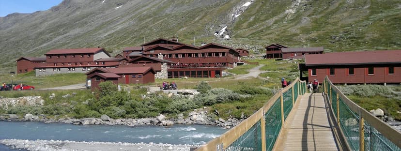 Wooden bridge over rushing river leading to Spiterstulen lodge by green mountain slope.