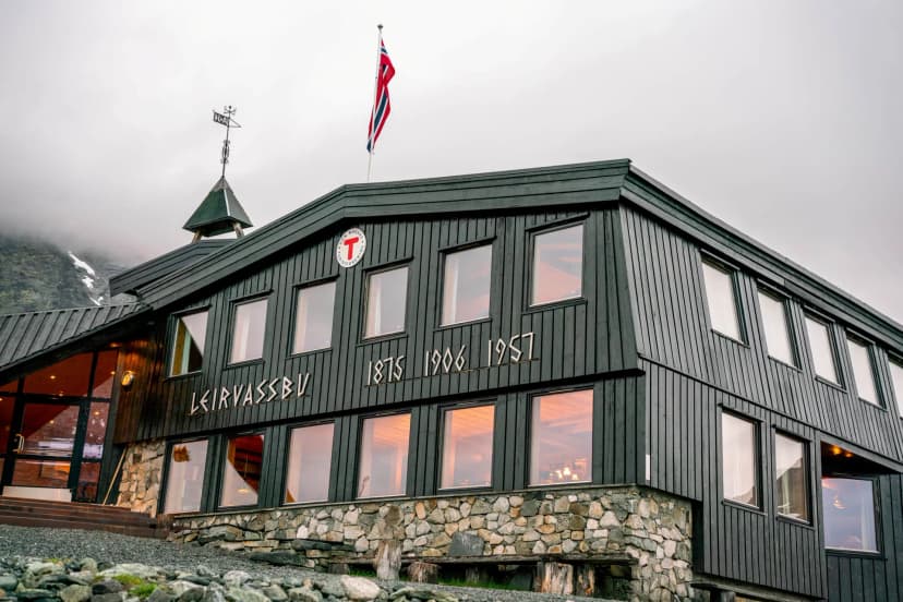 Leirvassbu mountain lodge with Norwegian flag under cloudy sky, stone foundation.