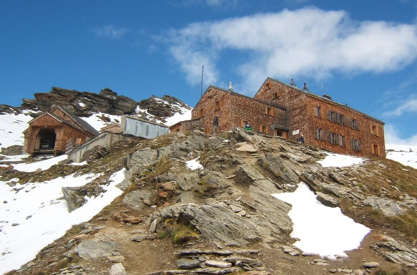 Defreggerhaus wooden mountain hut on rocky slope with patches of snow under blue sky.