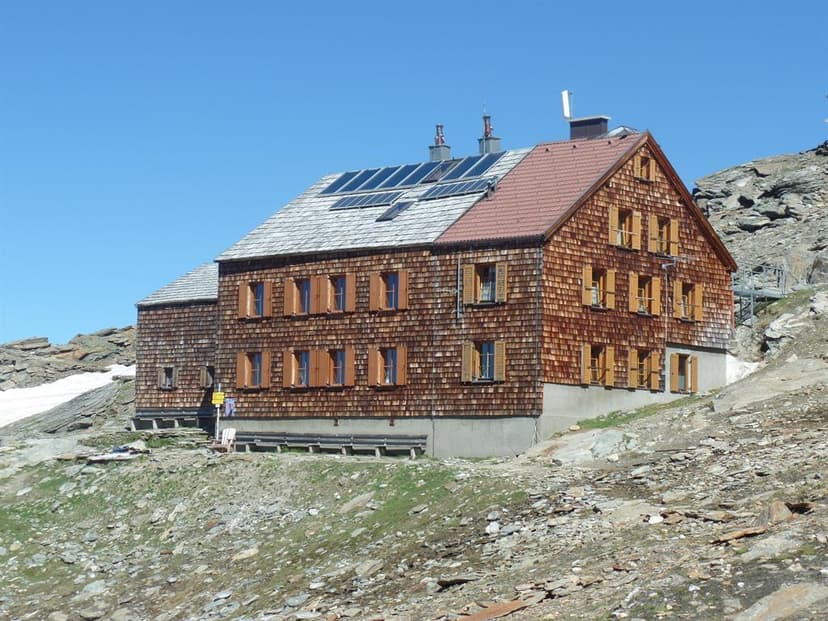 Wooden alpine hut with solar panels on the roof, set against a rocky slope under a clear blue sky.