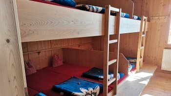 Wooden bunk beds with red mattresses and blue blankets inside a rustic mountain hut room.