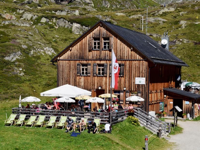 Johannishütte mountain hut with outdoor seating and deck chairs on grassy slope below rocky hill.