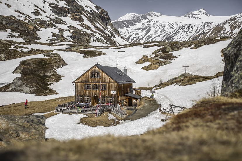Wooden mountain hut with outdoor seating surrounded by snow and high, snowy peaks, likely Johannishütte.
