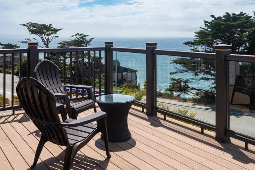 Balcony with two chairs and table overlooking ocean, coastal road, and cypress trees.