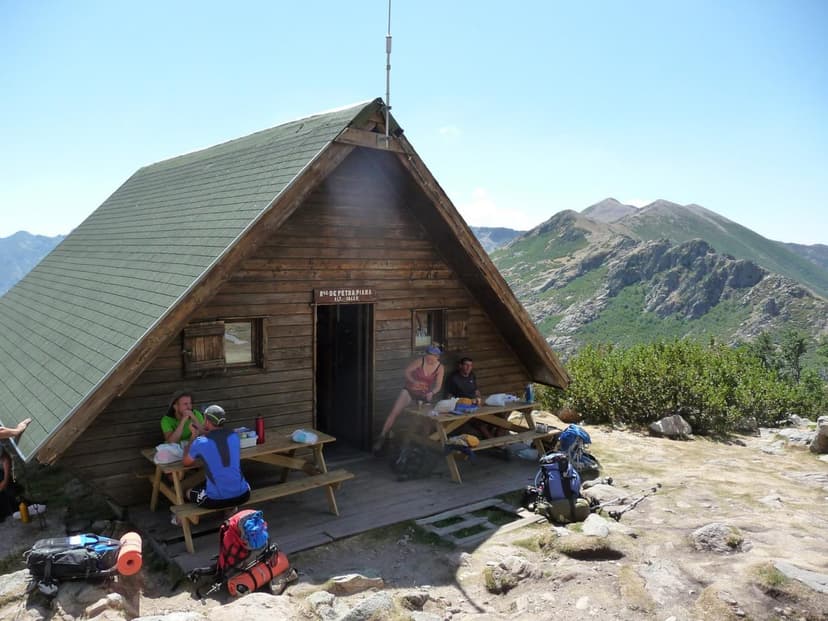 Hikers resting outside Refuge d'Ortu di u Piobbu with rugged mountains in background.