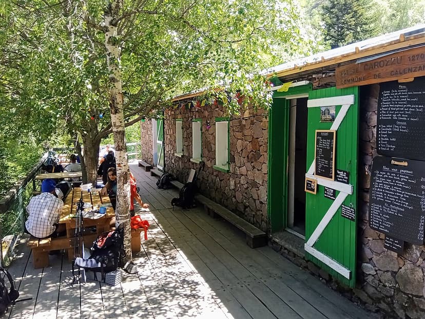 Refuge de Carrozzu stone building with green door and outdoor deck seating under trees.