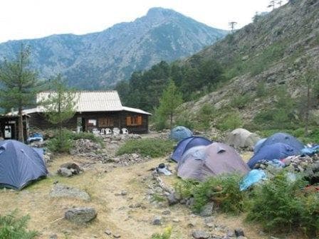 Tents pitched near a wooden mountain refuge with a large peak in the background.