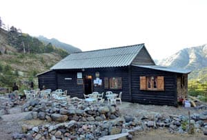 Dark wooden mountain refuge or cafe with outdoor seating surrounded by rocks and steep hillsides.