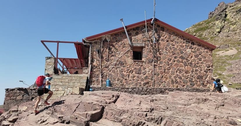 Hikers approaching Refuge de Ciottulu di i Mori stone mountain hut under clear blue sky.
