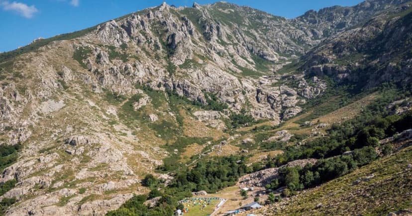 Rocky mountain valley with sparse vegetation and a small campsite below under a blue sky.