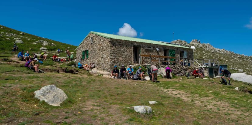 Hikers resting outside a stone mountain refuge with a green roof under a bright blue sky.