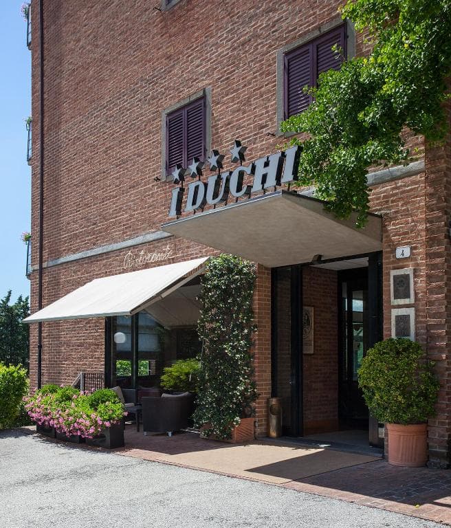 Hotel dei Duchi entrance with brick facade, awning, and potted flowers on a sunny day.