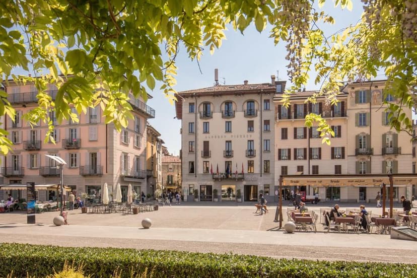 Albergo Firenze hotel facade on sunny piazza with outdoor cafe seating framed by green leaves.