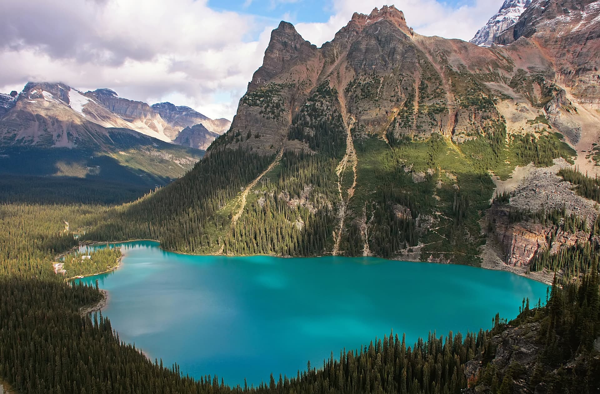 Turquoise alpine lake surrounded by dense forest and rugged, partially snow-capped mountains under cloudy sky.