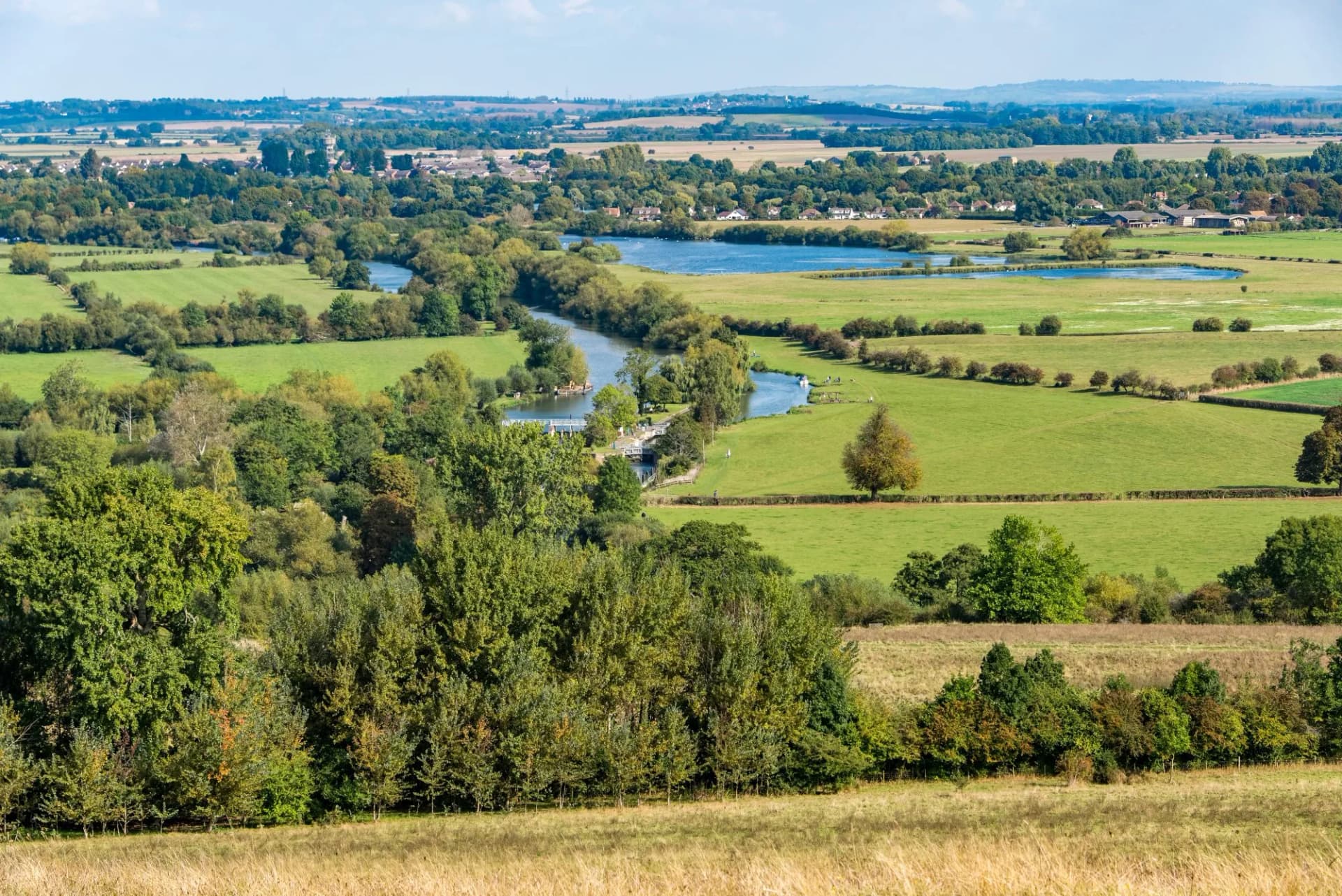 A sunlit rural scene with green fields and a river, seen from above. It is the River Thames seen from the hills known as the Wittenham Clumps, in Oxfordshire, United Kingdom.