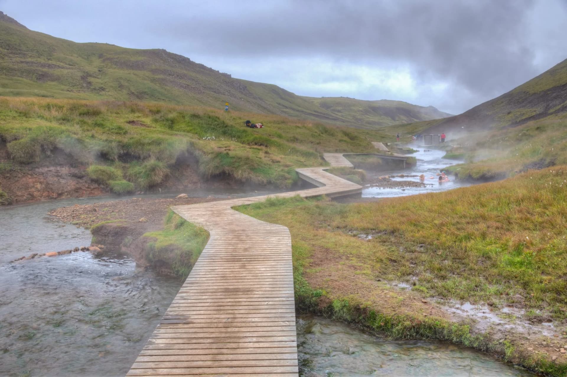 Wooden boardwalk along steaming river in Reykjadalur Valley, Iceland, with people bathing in hot spring.