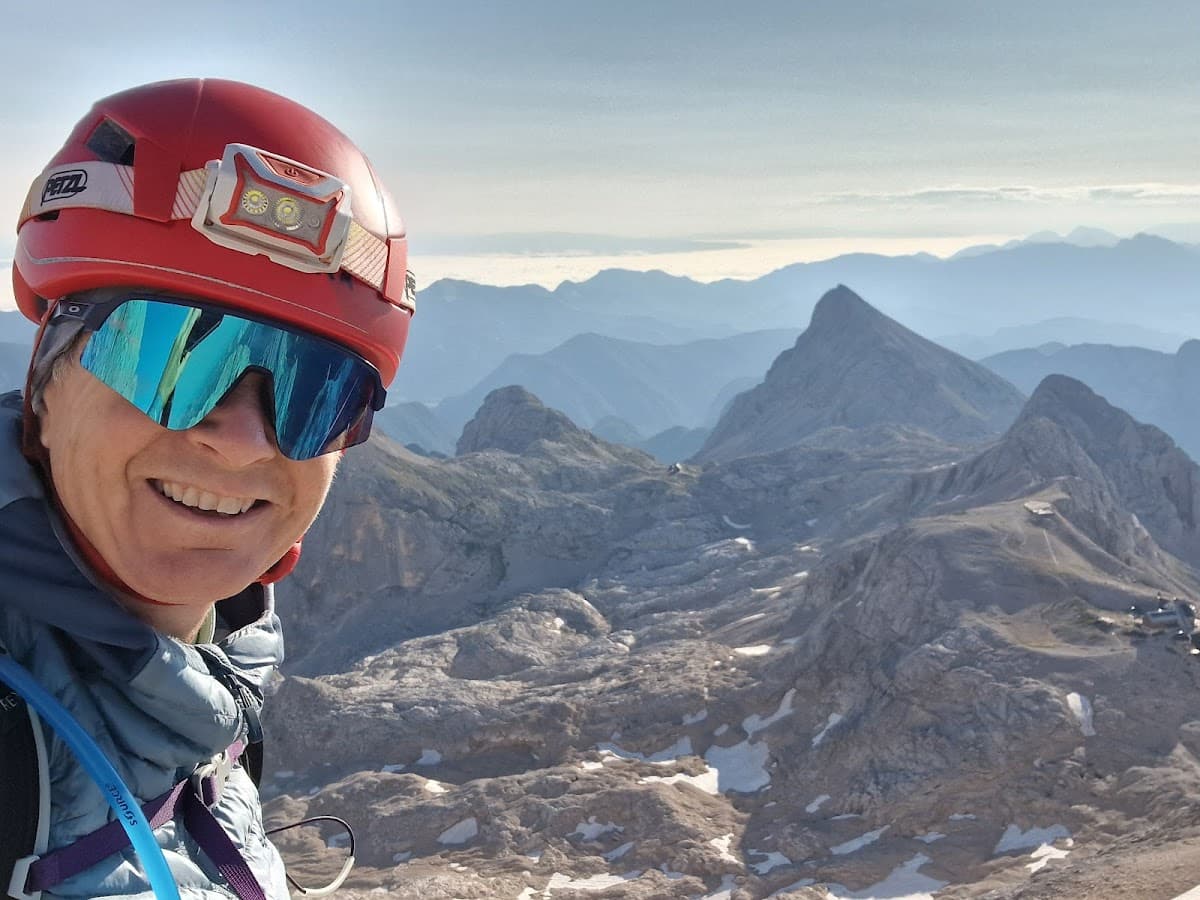 Hiker smiling in red helmet and sunglasses with rugged mountain peaks and snow patches below