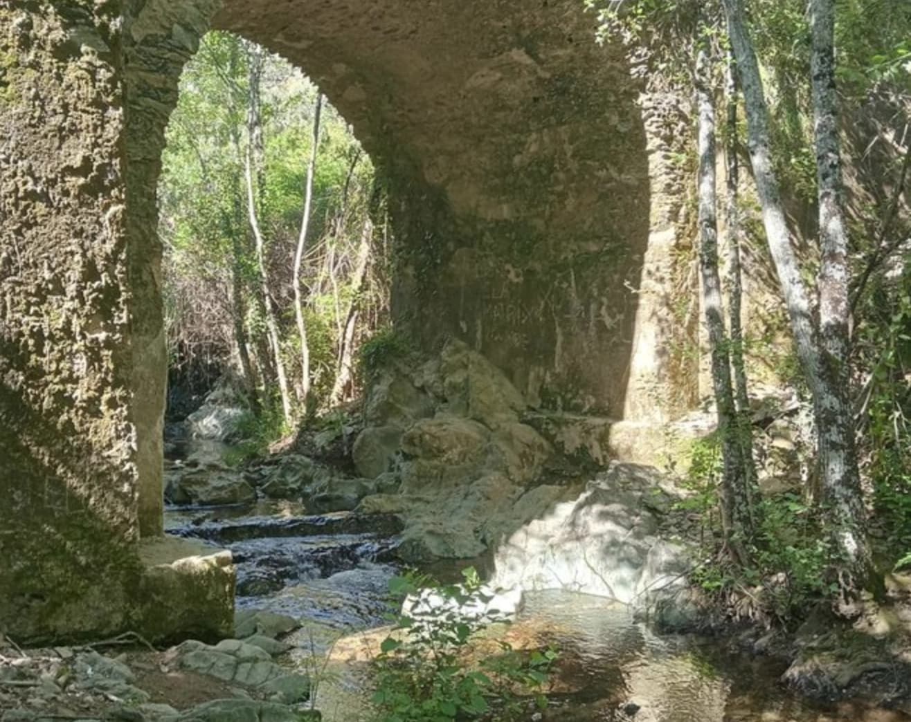 Stone arch bridge over a rocky stream in a sunlit forest setting, Guijarra.